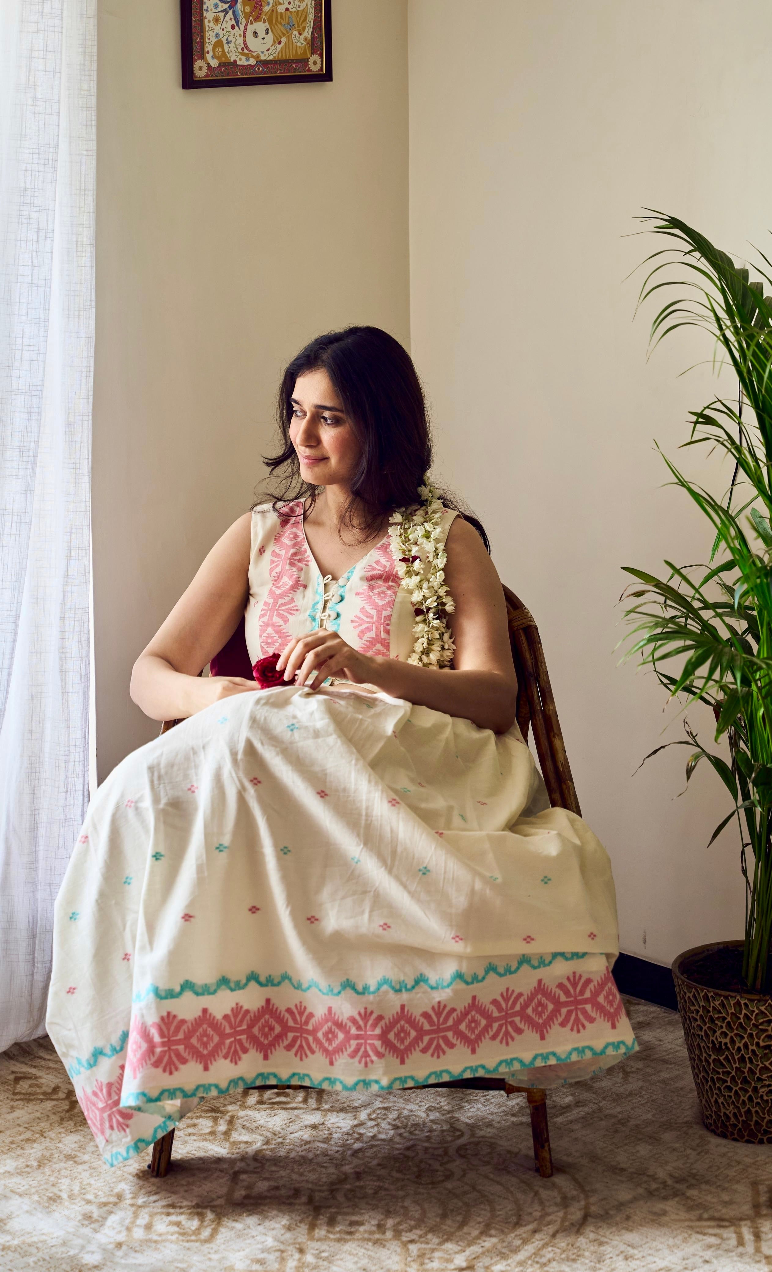 Woman in a traditional outfit sitting on a chair next to a potted plant indoors.